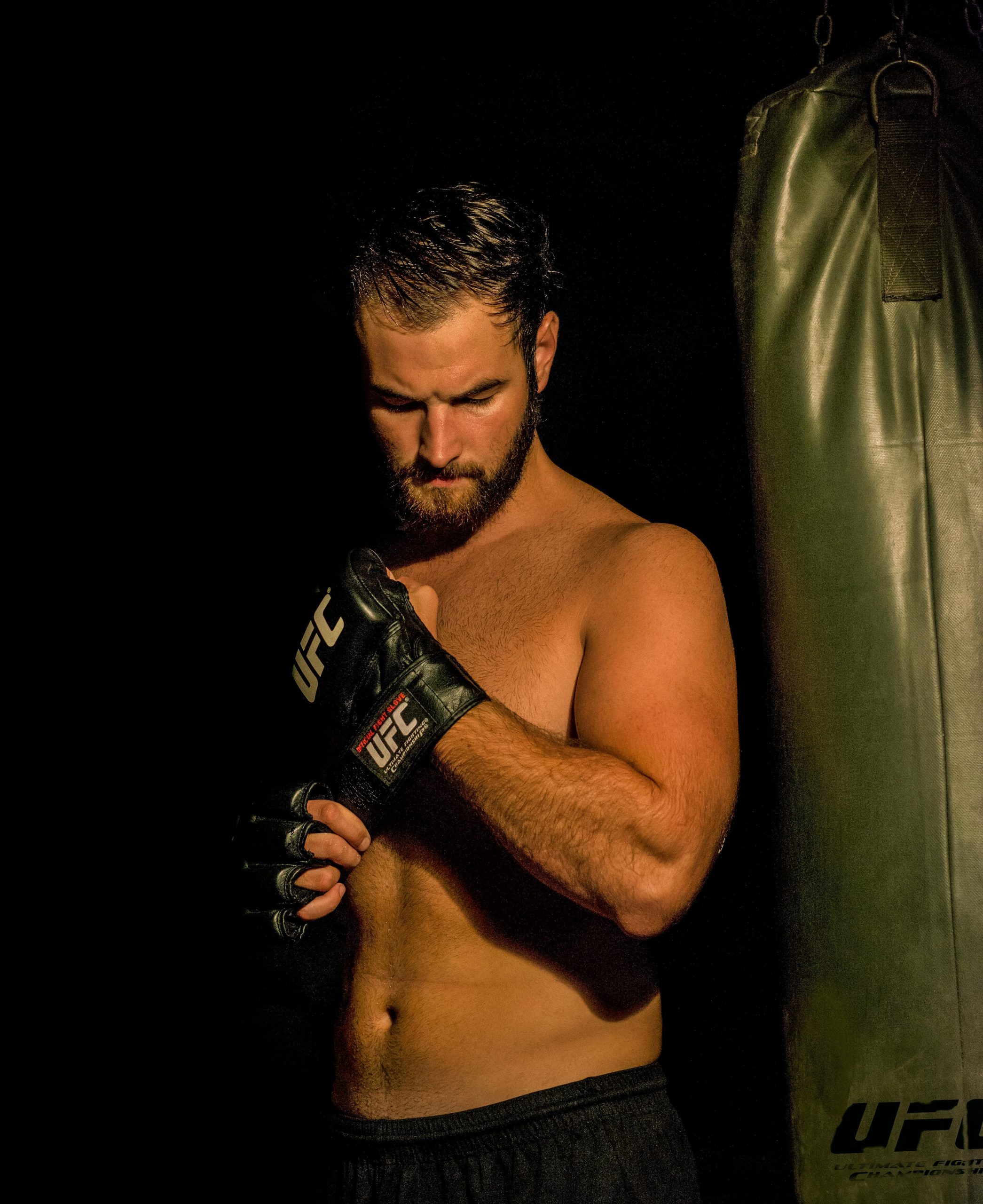 Shirtless male UFC fighter adjusting MMA gloves beside a heavy bag in dark gym lighting.