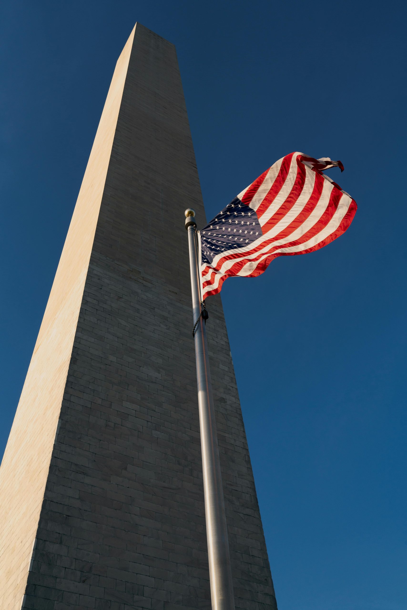 American flag waving beside the Washington Monument against a deep blue sky.
