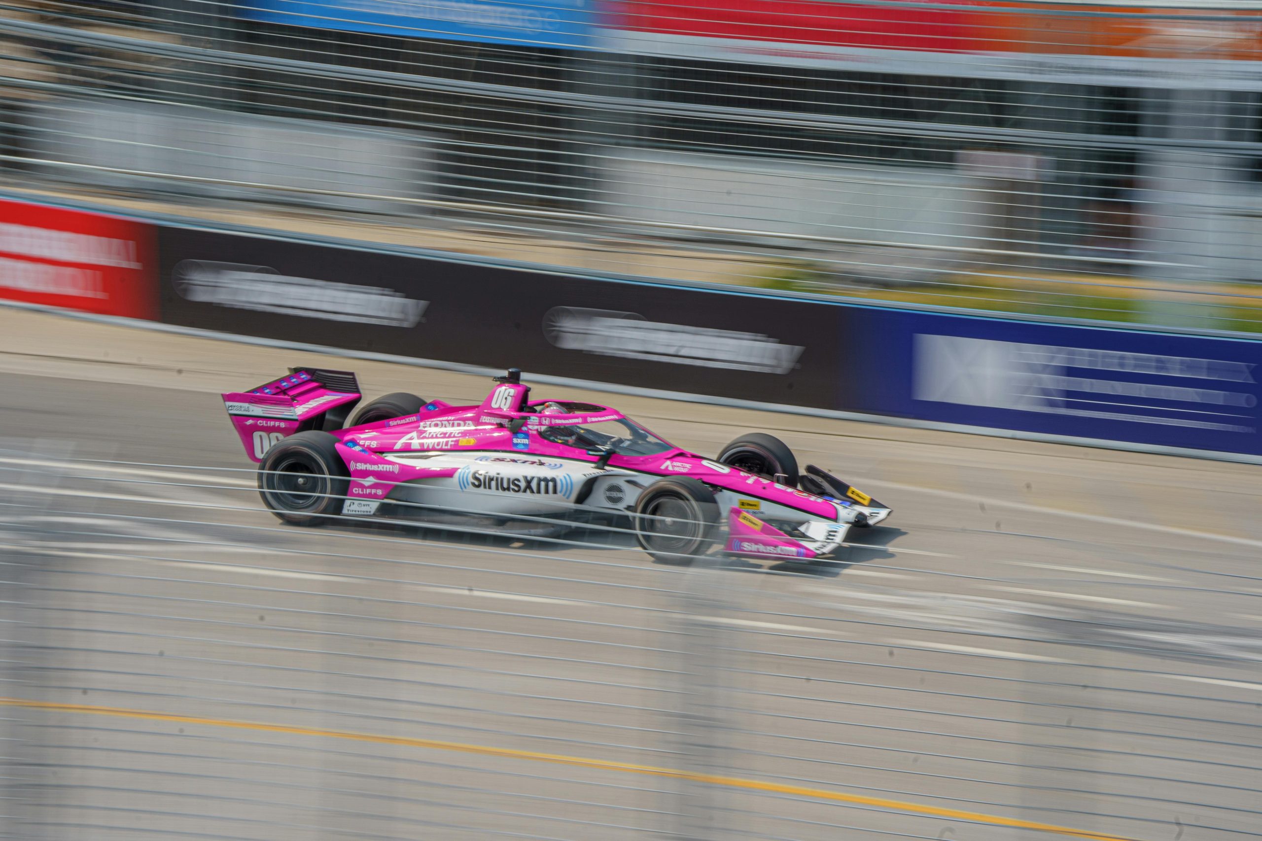 Pink and white SiriusXM-sponsored IndyCar racing at high speed on a street circuit.