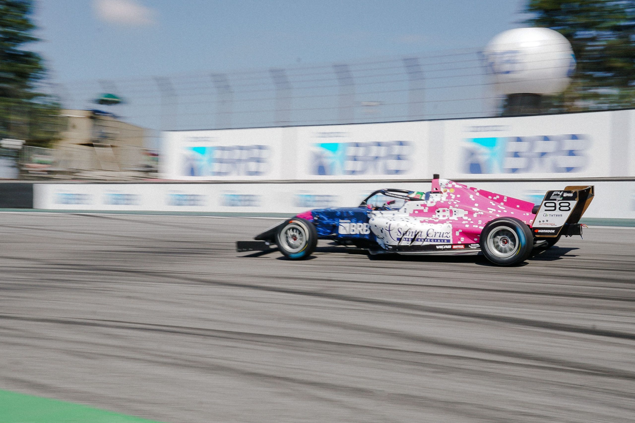 Pink, white, and blue Formula 4 race car speeding around a street circuit during a motorsport race.