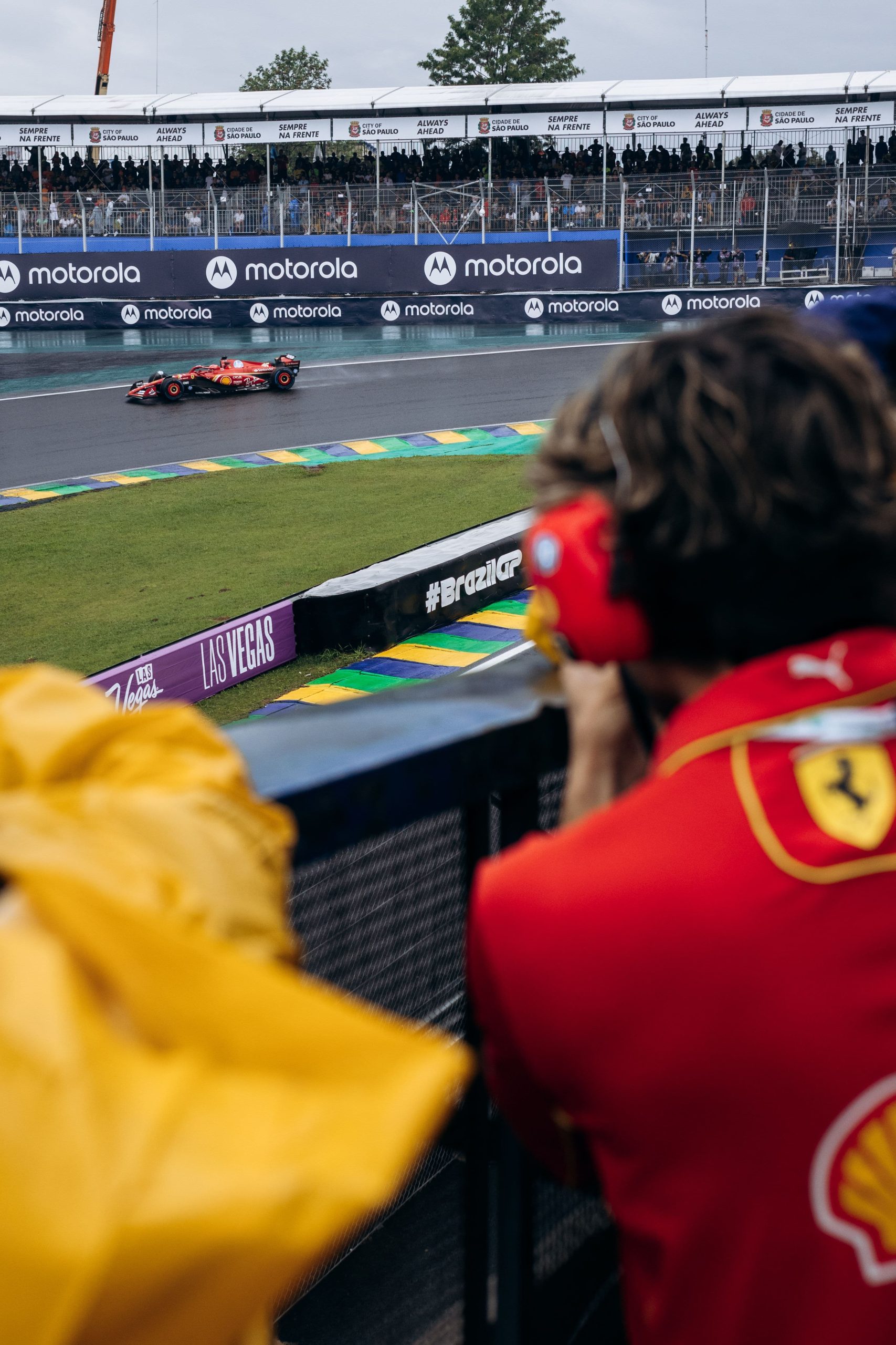 Spectator in Ferrari team gear photographing a red Formula 1 car rounding a curve on a rain-slicked track
